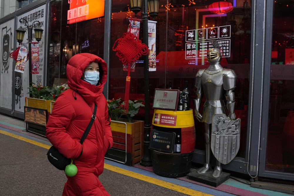 A woman wearing a mask to protect from the coronavirus walks past a coat of armour displayed outside a restaurant in Beijing, China, Friday, Jan. 21, 2022.